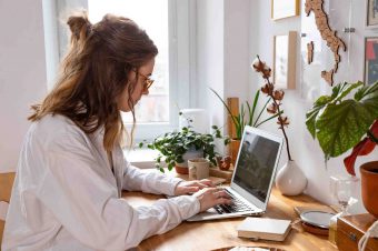 Young woman freelancer/designer working on computer from home office during self-isolation due to coronavirus. Cozy workplace surrounded by plants. Remote work, Telecommuting, Distance job.